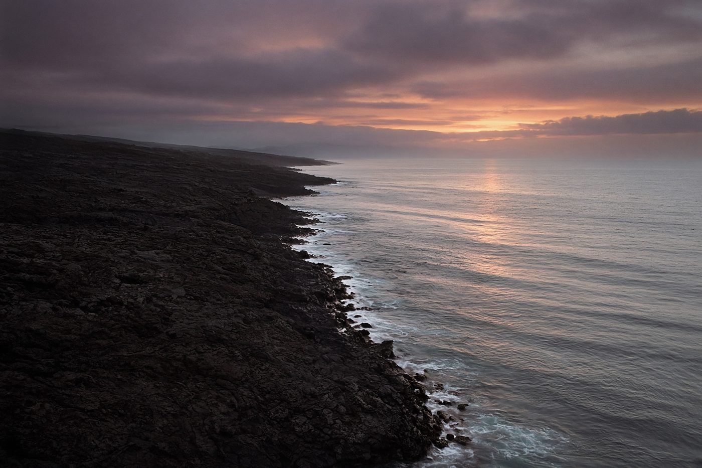 Hawaiian volcanic coastline at dusk, dark lava meeting a pale ocean.