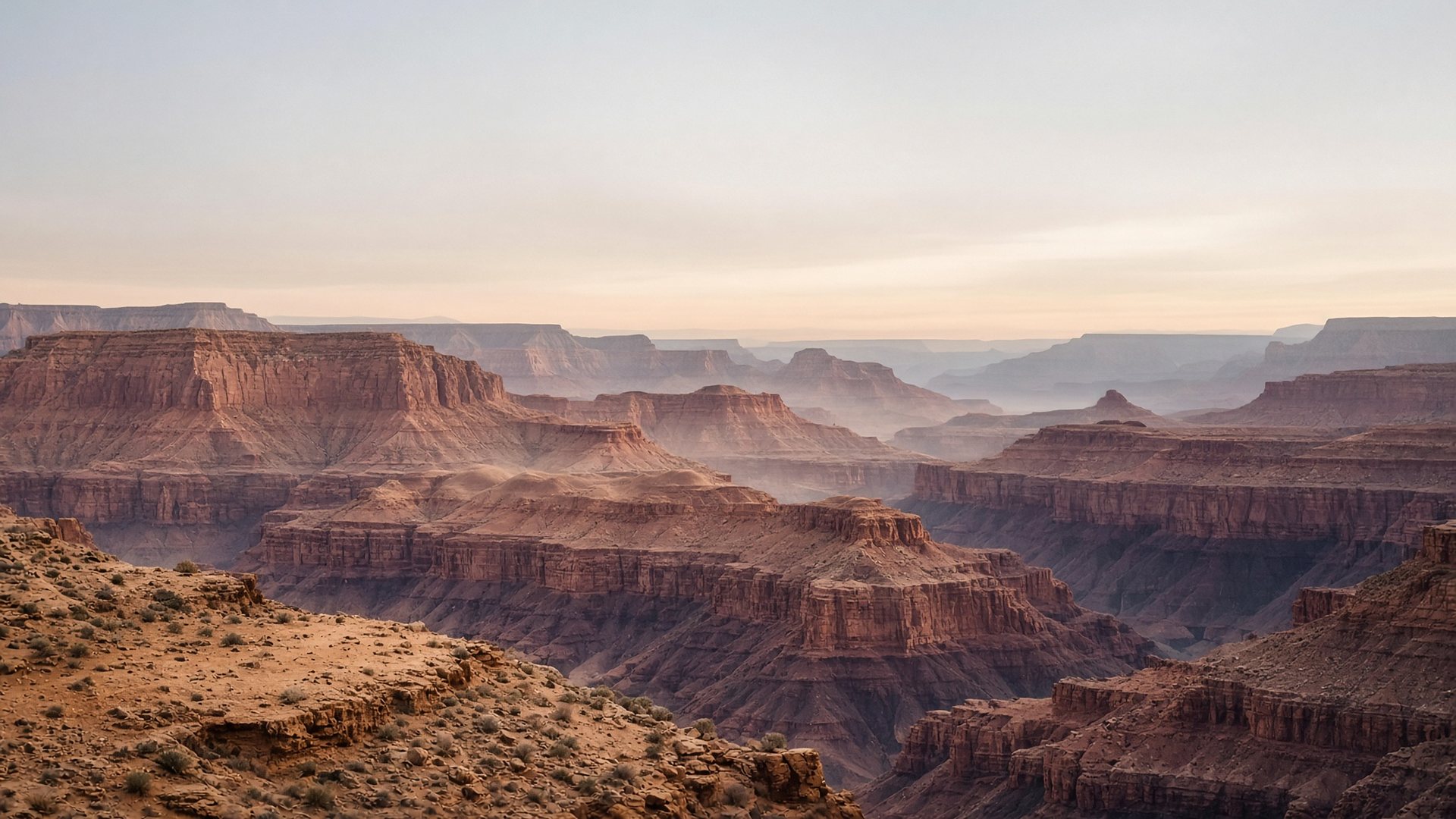 Layered canyon country at first light, receding into thin weather under a pale sky.